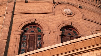A close-up view of an architectural structure made of stone, featuring arched windows with intricately designed stained glass. The stonework includes decorative elements and patterns, highlighting the craftsmanship involved in the building's construction.