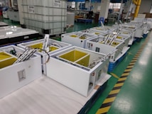 A factory setting with multiple white rectangular machines lined up on a green-tiled floor. Each machine has open compartments with visible wiring and metal components, surrounded by several large tanks and various industrial equipment.