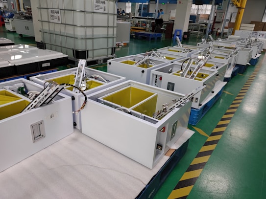 A factory setting with multiple white rectangular machines lined up on a green-tiled floor. Each machine has open compartments with visible wiring and metal components, surrounded by several large tanks and various industrial equipment.