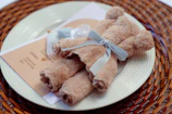A beige-colored towel is folded and arranged to resemble a teddy bear, tied with a light blue ribbon. It is placed on a white plate, which is set on a woven placemat. A card with printed text is partially visible beneath the towel.