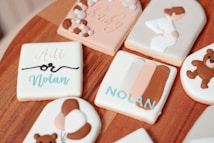 Decorative cookies are arranged on a wooden surface, featuring pastel colors and thematic designs related to babies and pregnancy. One cookie has 'Adli or Nolan' written on it in script, suggesting a gender reveal theme. Another cookie features an illustration of a pregnant woman. Additional designs include teddy bears and a card with color swatches.