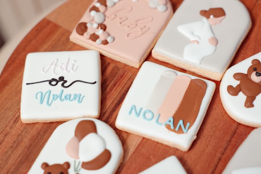 Decorative cookies are arranged on a wooden surface, featuring pastel colors and thematic designs related to babies and pregnancy. One cookie has 'Adli or Nolan' written on it in script, suggesting a gender reveal theme. Another cookie features an illustration of a pregnant woman. Additional designs include teddy bears and a card with color swatches.