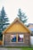 A small wooden building with a triangular roof has a poster displayed in one of its windows. The poster features a group of people wearing helmets, suggesting an adventure or outdoor activity. The structure is made of logs and has a rustic appearance. Tall evergreen trees and another building are visible in the background.
