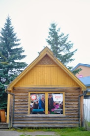 A small wooden building with a triangular roof has a poster displayed in one of its windows. The poster features a group of people wearing helmets, suggesting an adventure or outdoor activity. The structure is made of logs and has a rustic appearance. Tall evergreen trees and another building are visible in the background.