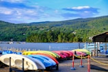A scenic waterfront with an array of colorful kayaks and paddleboards neatly lined up on a sandy shore. In the background, a tranquil lake is bordered by a lush green forest and rolling hills under a partly cloudy sky. A few boats float on the water, contributing to the idyllic setting.