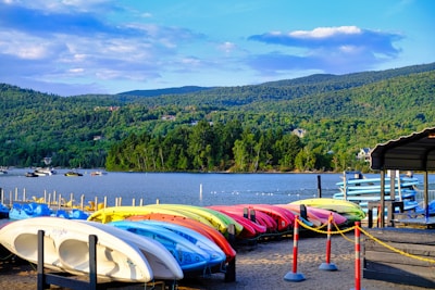 A scenic waterfront with an array of colorful kayaks and paddleboards neatly lined up on a sandy shore. In the background, a tranquil lake is bordered by a lush green forest and rolling hills under a partly cloudy sky. A few boats float on the water, contributing to the idyllic setting.