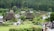 A serene village scene with traditional thatched-roof houses surrounded by lush greenery and tall trees. People are walking along a path through the village, suggesting a peaceful and leisurely atmosphere.