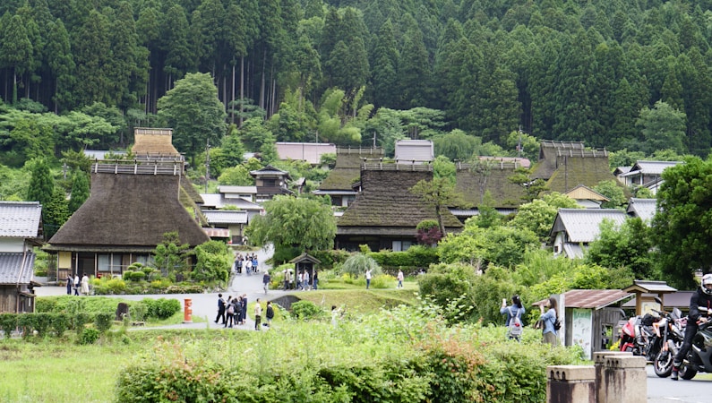 A serene village scene with traditional thatched-roof houses surrounded by lush greenery and tall trees. People are walking along a path through the village, suggesting a peaceful and leisurely atmosphere.