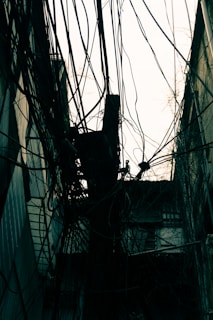 A complex web of tangled wires hangs between buildings in an urban setting. The wires are intertwined around a pole, creating a chaotic and dense network. The background features the outlines of old, worn buildings that contribute to the gritty city atmosphere.