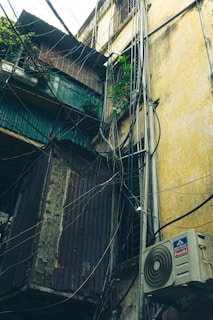 An exterior view of an old building with numerous electrical wires crisscrossing and a corrugated metal structure attached. An air conditioning unit is mounted on the wall, and green plants are visible in a small space. The building walls are weathered, with a faded yellow and brown color.