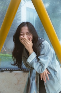 A woman with long dark hair is laughing and covering her mouth with her hand. She is wearing a denim jacket and is leaning slightly forward, with a natural background blurred behind her.