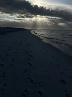 A sunlit beach with footprints leading toward a glowing horizon under a clear blue sky.