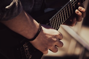 A person is playing an electric guitar, focusing on one hand pressing the guitar strings while the other hand plucks them. The lighting casts dramatic shadows, emphasizing the textures of the guitar and the person's hand.