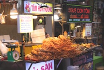 The image depicts a street food stall selling grilled lobsters. The stall features various signs in Vietnamese advertising prices and menu options. There is a large pile of lobsters prominently displayed at the front of the stall. The environment appears to be busy with multiple light sources illuminating the area. Some kitchen equipment and scales are visible.