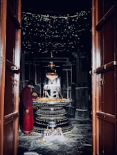 A dimly lit temple room containing a large, decorated lingam at the center. A person dressed in traditional attire stands nearby, possibly engaged in religious activity. The lingam is adorned with garlands and inscriptions, with small decorative items placed around it. Wooden doors frame the entrance to the room.