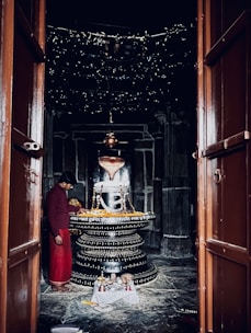 A dimly lit temple room containing a large, decorated lingam at the center. A person dressed in traditional attire stands nearby, possibly engaged in religious activity. The lingam is adorned with garlands and inscriptions, with small decorative items placed around it. Wooden doors frame the entrance to the room.