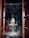 A dimly lit temple room containing a large, decorated lingam at the center. A person dressed in traditional attire stands nearby, possibly engaged in religious activity. The lingam is adorned with garlands and inscriptions, with small decorative items placed around it. Wooden doors frame the entrance to the room.