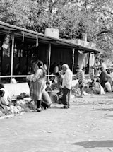 A busy outdoor market scene where people are engaged in buying and selling goods. There are several individuals, including adults and children, interacting and crouching beside various produce displayed on the ground. The market appears to be bustling with activity under a canopy, and there are leaves scattered on the pavement.