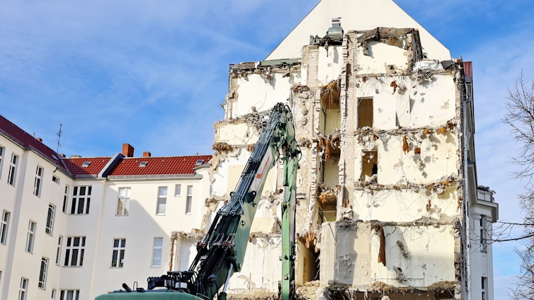 A partially demolished building with exposed bricks and rubble, surrounded by intact buildings with red roofs. A large excavator arm is visible, indicating ongoing demolition work under a clear blue sky.