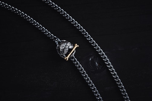 A dark metal chain necklace with a pendant resembling a lion's head clenching a gold-colored bone. The chain and pendant rest on a black surface.