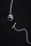 A silver chain with a prominent pendant at one end shaped like a detailed dog head and a small gold bone-shaped toggle clasp on a dark background. The overall composition is minimalistic and artistic, highlighting the metallic textures and intricate designs of the jewelry piece.