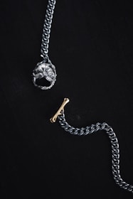 A silver chain with a prominent pendant at one end shaped like a detailed dog head and a small gold bone-shaped toggle clasp on a dark background. The overall composition is minimalistic and artistic, highlighting the metallic textures and intricate designs of the jewelry piece.