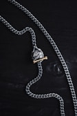 A silver chain necklace featuring a pendant shaped like a skull is laid out on a dark wooden surface. The pendant has a brass bone placed beneath the skull, adding contrast to the metallic tones.