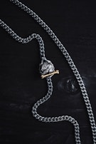 A silver chain necklace featuring a pendant shaped like a skull is laid out on a dark wooden surface. The pendant has a brass bone placed beneath the skull, adding contrast to the metallic tones.