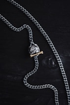 A silver chain necklace featuring a pendant shaped like a skull is laid out on a dark wooden surface. The pendant has a brass bone placed beneath the skull, adding contrast to the metallic tones.