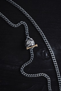 A silver chain necklace featuring a pendant shaped like a skull is laid out on a dark wooden surface. The pendant has a brass bone placed beneath the skull, adding contrast to the metallic tones.