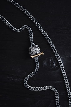 A silver chain necklace featuring a pendant shaped like a skull is laid out on a dark wooden surface. The pendant has a brass bone placed beneath the skull, adding contrast to the metallic tones.