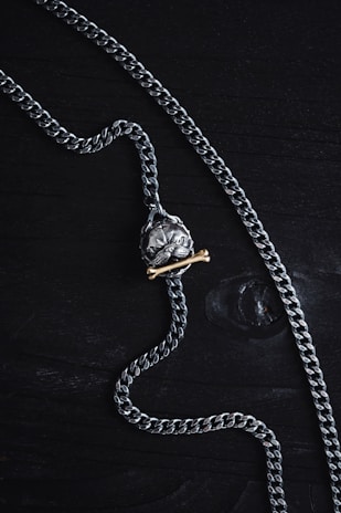 A silver chain necklace featuring a pendant shaped like a skull is laid out on a dark wooden surface. The pendant has a brass bone placed beneath the skull, adding contrast to the metallic tones.