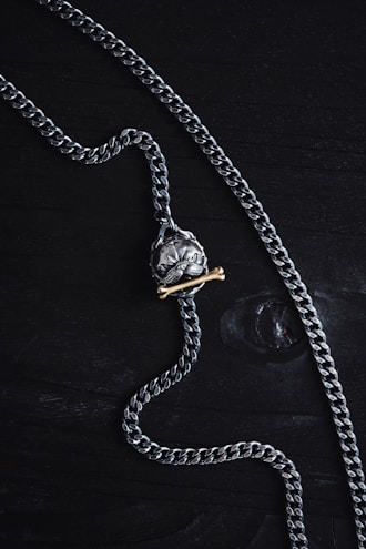A silver chain necklace featuring a pendant shaped like a skull is laid out on a dark wooden surface. The pendant has a brass bone placed beneath the skull, adding contrast to the metallic tones.