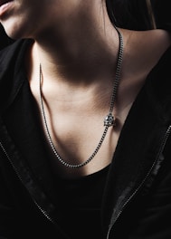 A person wearing a black jacket and a metallic chain necklace, featuring a skull pendant. The lighting is dramatic, casting shadows that highlight the texture of the skin and clothing.