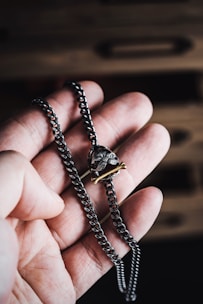 A hand holding a chunky metal chain necklace with a pirate-themed pendant featuring a skull and crossed bones. The background is blurred wood surface, adding contrast to the focus on the jewelry.