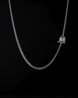 A delicate silver chain with a heart-shaped crystal pendant is suspended against a dark background. The pendant appears to be attached to a small golden bar.