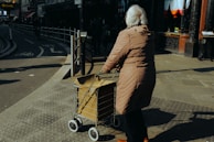 An elderly person with white hair, wearing a long beige coat, is pushing a shopping cart on a city sidewalk. The scene takes place during daytime in an urban area, with a bicycle lane and buildings in the background. The lighting suggests a bright, sunny day.