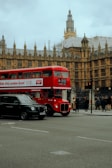 A classic red London double-decker bus is passing by an iconic historical building with intricate Gothic architecture. Nearby, a black taxi is on the road, while several pedestrians are walking along the sidewalk. The sky is overcast, adding a muted tone to the scene.