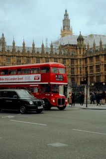 A classic red London double-decker bus is passing by an iconic historical building with intricate Gothic architecture. Nearby, a black taxi is on the road, while several pedestrians are walking along the sidewalk. The sky is overcast, adding a muted tone to the scene.
