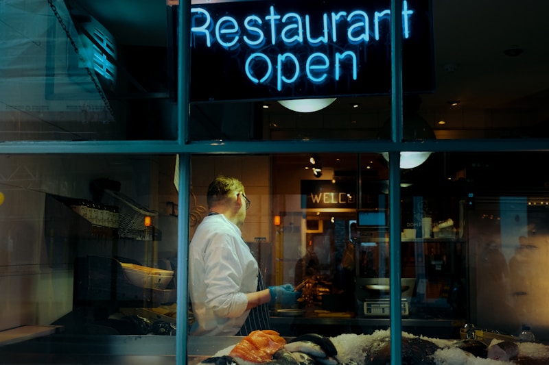 A view through a window into a restaurant, where a person wearing a white shirt is standing behind a counter with various seafood items on ice. A neon sign reading 'Restaurant Open' is prominently displayed above the window. The interior is warmly lit, with reflections visible on the glass.