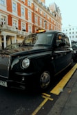 A classic black taxi is parked on the side of a street with a red brick building in the background. The taxi has a distinct shape and design typical of British cabs and is positioned next to a yellow line.