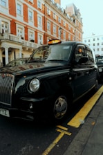 A classic black taxi is parked on the side of a street with a red brick building in the background. The taxi has a distinct shape and design typical of British cabs and is positioned next to a yellow line.