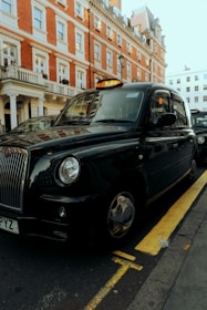 A classic black taxi is parked on the side of a street with a red brick building in the background. The taxi has a distinct shape and design typical of British cabs and is positioned next to a yellow line.