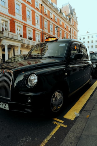 A classic black taxi is parked on the side of a street with a red brick building in the background. The taxi has a distinct shape and design typical of British cabs and is positioned next to a yellow line.