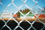 A chain-link fence is in the foreground, providing a grid-like pattern through which a train yard is visible. The train yard contains multiple colorful shipping containers stacked on flatbed train cars. Several containers have 'MAERSK' branding, with a prominent blue container and others in red and green. Trees line the background against a clear blue sky.