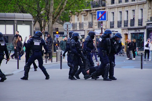 A group of police officers in riot gear are detaining a person on a city street. The officers are wearing helmets and protective clothing, and they appear to be escorting the individual away. Several onlookers and passersby are visible in the background, walking on the sidewalk and observing the scene. Urban buildings and signage can also be seen around the area.