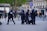 A group of police officers in riot gear are detaining a person on a city street. The officers are wearing helmets and protective clothing, and they appear to be escorting the individual away. Several onlookers and passersby are visible in the background, walking on the sidewalk and observing the scene. Urban buildings and signage can also be seen around the area.