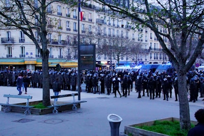 A large gathering of riot police is positioned in a street, with police vans parked nearby. The officers are wearing helmets and shields, and stand in front of a multi-story building with balconies. The area includes some trees, benches, and bystanders walking nearby.
