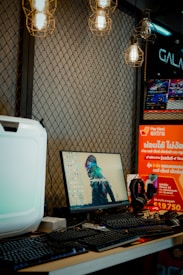 A desk setup featuring a monitor displaying a cyberpunk artwork, a keyboard, and headphones. The background is a mesh wall adorned with hanging industrial-style light bulbs. To the right, there are promotional posters with bright colors and text in a foreign language.