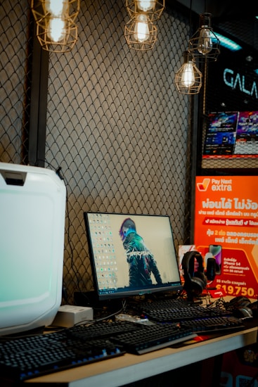 A desk setup featuring a monitor displaying a cyberpunk artwork, a keyboard, and headphones. The background is a mesh wall adorned with hanging industrial-style light bulbs. To the right, there are promotional posters with bright colors and text in a foreign language.